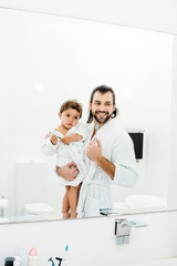 Fototapeta premium Dad and toddler son looking in mirror at home in white bathroom
