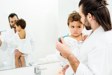 Fototapeta premium Dad looking at son and showing tothbrush with toothpaste in white bathroom