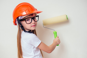cute and little girl in a construction helmet hard hat and glasses holding paint roller paints a...