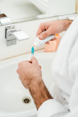 cropped view of male hands adding toothpaste on toothbrush in bathroom