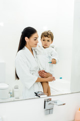 mother and son in white bathrobes brushing teeth together in bathroom