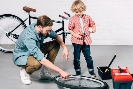 male repairman fixing bicycle wheel with pliers while his little son standing near in workshop