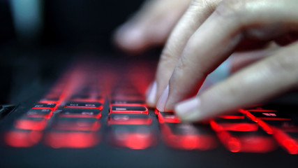 Teenage Hacker Girl Attacks Corporate Servers in Dark, Typing on Red Lit Laptop Keyboard. Room is...