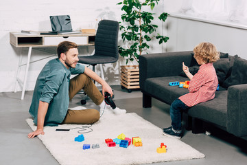 Naklejka premium child sitting on sofa while his father holding smartphone with headphones on floor at home