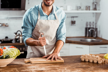 cropped shot of smiling young man in apron holding knife and hand on chopping board in kitchen