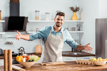 cheerful young man in apron with open arms smiling at camera in kitchen