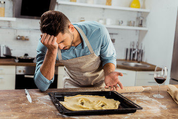 upset young man in apron leaning at table with spoiled dough on baking tray
