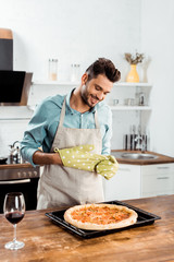 smiling young man in apron and potholders looking at fresh homemade pizza on baking tray