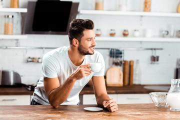 smiling young man holding cup of coffee and leaning at kitchen table at morning - Powered by Adobe