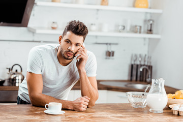 young man leaning at kitchen table and talking by smartphone at morning