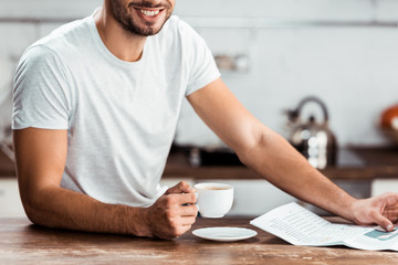 cropped shot of smiling young man holding cup of coffee and newspaper at morning