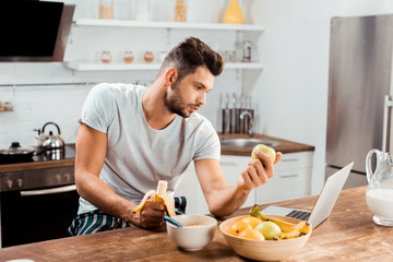 Fototapeta na wymiar young man in pajamas holding fruits and using laptop at morning