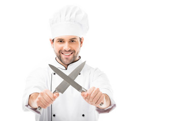 smiling young chef holding cutting knives in shape of x and looking at camera isolated on white