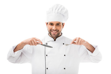 happy young chef holding fork and knife and looking at camera isolated on white