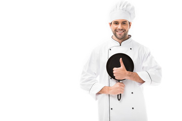 smiling young chef holding frying pan and showing thumb up isolated on white