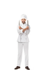 Fototapeta na wymiar handsome young chef holding knives in crossed arms and smiling at camera isolated on white
