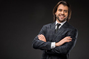 handsome smiling young businessman standing with crossed arms and looking away isolated on black