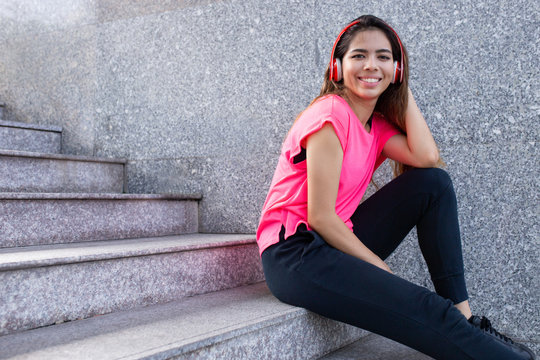 Portrait of happy teenage girl sitting on stairs in headphones. Young Latin American woman listening to music outdoors. Music and youth culture concept
