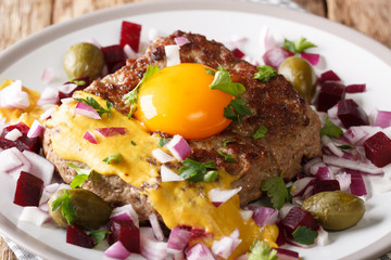 Pariserbof is a patty of lean ground beef on a slice of white bread, panfried and served with raw onion, pickled beets, capers and a raw egg yolk closeup on a plate. Horizontal