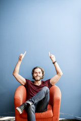 Portrait of a young caucasian bearded man with long hair showing with hands on the colorful background sitting on the chair. Image with copy space