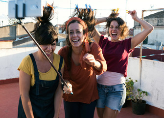 Happily laughing beautiful Hispanic female friends standing on roof terrace with hair attached to washing line with clothes-pegs and taking selfie on smartphone with stick on sunny summer day 