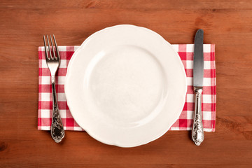 An overhead photo of a white plate with a fork and a knife on a gingham cloth on a dark rustic wooden background with copy space