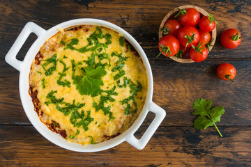 Ground beef baked with tomato, bell pepper, onion and rice and topped with grated cheese and fresh parsley in white casserole on wooden rustic table