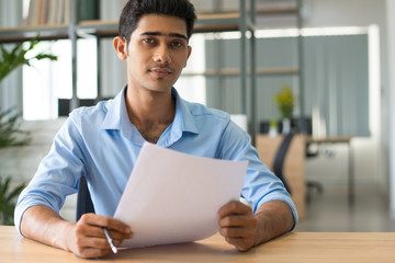 Content confident handsome Indian businessman analyzing data while examining report in office. Serious young business expert working with document. Paperwork concept