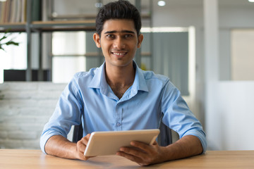 Positive confident Indian man in blue shirt using tablet in modern office. Smiling handsome young employee sitting at table and analyzing data. Businessman concept