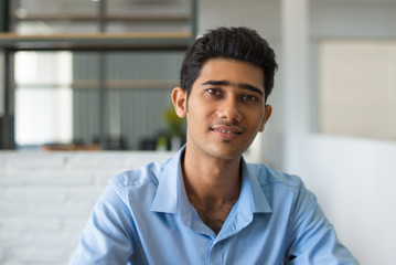 Smiling young Indian businessman in blue shirt looking at camera. Positive successful male entrepreneur sitting in modern office. Success concept
