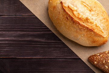 Simple minimalistic bread background. A white wheat loaf with a crispy fragrant crust and seed bun lie on parchment over dark natural pine planks of the table. Parchment divides the frame diagonally.