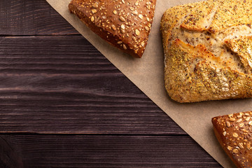 Bread background. Two rye triangles and one wheat square delicious freshly baked buns with seeds and crispy fragrant crust lie on parchment over dark natural pine planks of the table. Copy space.