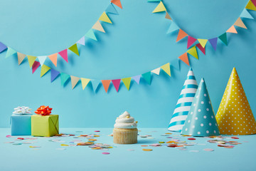  Tasty cupcake, party hats, confetti and gifts on blue background with colorful bunting