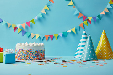 Tasty cake with sugar sprinkles,party hats and gifts on blue background with colorful bunting - Powered by Adobe