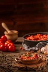 Delicious meat and tomato meal served in ceramic dish with handles and fresh vegetables and ingredients on wooden table on dark background