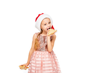 happy little child in santa hat eating ginger cookies and looking at camera isolated on white