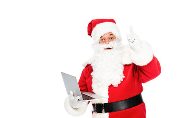 santa claus holding laptop and looking at camera while pointing up isolated on white