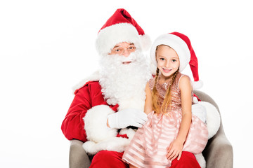 Fototapeta na wymiar happy santa claus and little child sitting in armchair together and looking at camera isolated on white