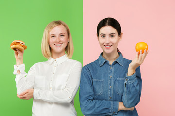 Diet. Dieting concept. Healthy useful food. Beautiful young women choosing between fruits and unhelathy fast food at studio. Human emotions and comparison concepts