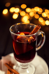 closeup glass of mulled wine with orange and cinnamon on dark black background, on white plate, Christmas lights