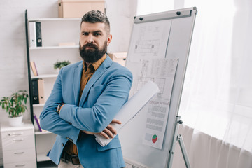 serious adult male architect in blue formal wear looking at camera, holding blueprint, using flip chart and working on project in office