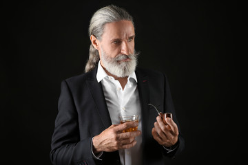 Elderly businessman with glass of whiskey and pipe on dark background