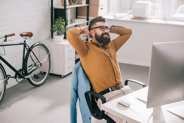 smiling bearded adult businessman in earphones with hands behind head sitting and relaxing at computer desk in office