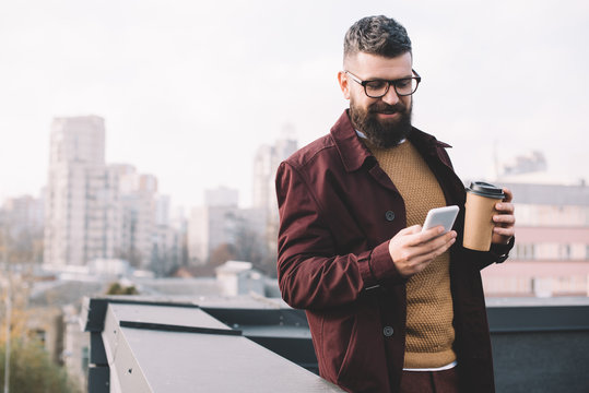 stylish adult man in glasses holding coffee to go and using smartphone on rooftop