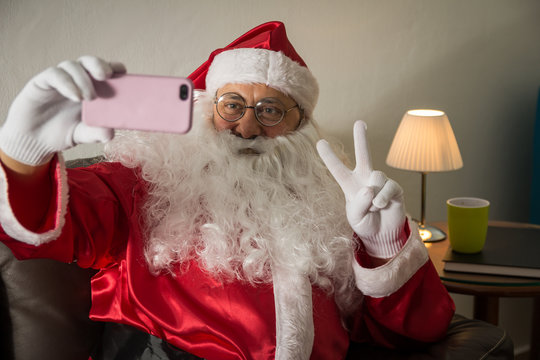 Front view of Santa Claus sitting in sofa at home Using cell phone for communication and leisure. Taking a photograph of himseelf, selfie.