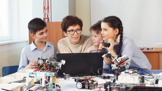 Curious clever pupils with the help of their female teacher doing a group project programming homemade robot using laptops on extracurricular classes