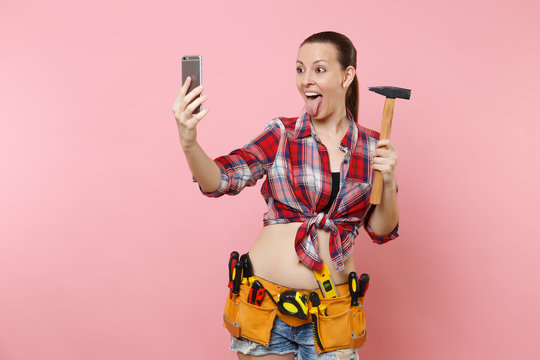 Excited fun handyman woman in plaid shirt, denim shorts, kit tools belt full of instruments, hammer doing selfie on mobile phone isolated on pink background. Female in male work. Renovation concept