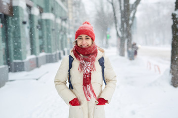 Cheerful smiling woman in white down jacket and red cap, scarf and mittens walking on the snowy street after blizzard in city. winter city after blizzards and snowfall.