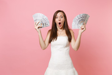 Shocked bride woman with opened mouth in white wedding dress holding bundle lots of dollars, cash money isolated on pink pastel background. Organization of wedding celebration concept. Copy space.