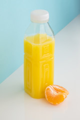 Orange drink and wedge of tangerine on white and blue background. Minimalistic image of citrus juice bottle and fruit at sparse bright environment.